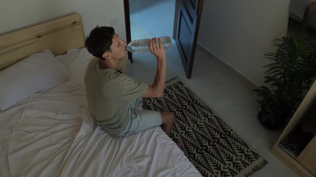 Hot climate lifestyle as man drinks water while sitting on bed during intense daytime heat, showing healthy hydration habits and everyday adaptation to prolonged hot weather at home,
