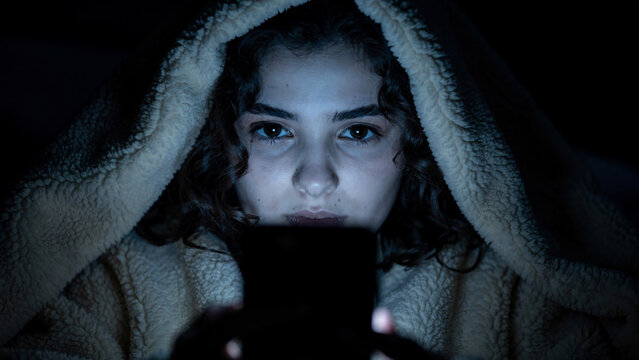 Young woman with curly hair under a fuzzy hood illuminated by a smartphone screen, close-up, concept for screen time, digital addiction and modern communication