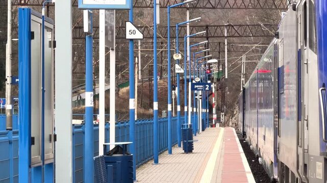A passenger train arrives at a modern railway station platform with blue structural poles and infrastructure reflecting daily commuter transit journeys within a scenic mountain area