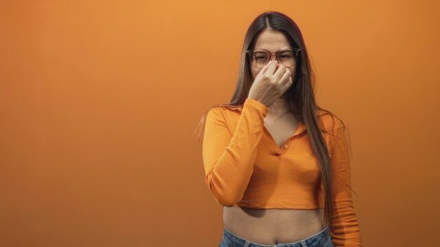 Young hispanic woman pinches nose with hand, wearing glasses and orange cropped top in a studio; disgust odor problem.