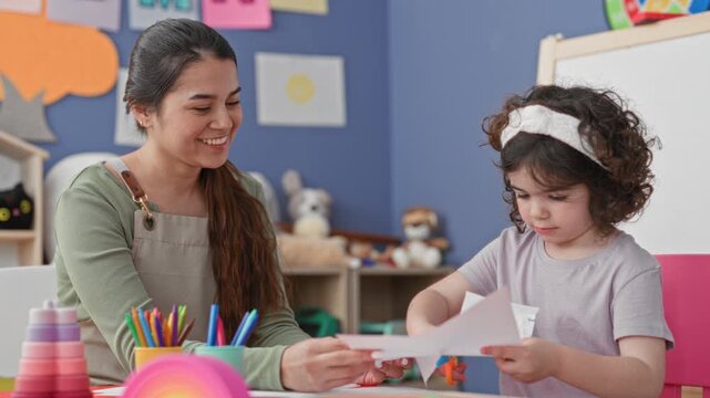 Teacher helps girl cut paper, holding sheet and scissors at table in building; nurturing learning calm.