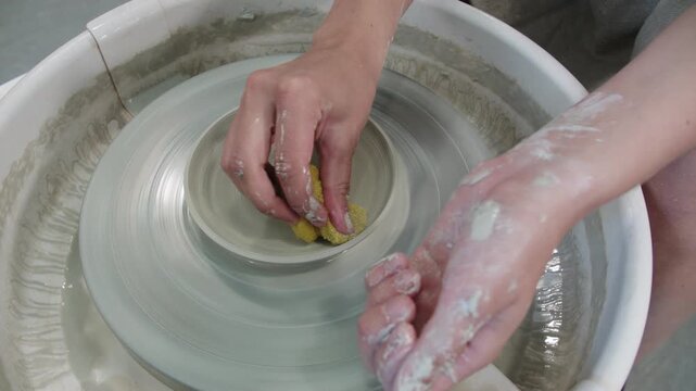Potter smoothes edges of plate and absorption of excess water of product during work on circle with synthetic sponge. Final stage of work on ceramic product. Handicraft workshop with potters wheel