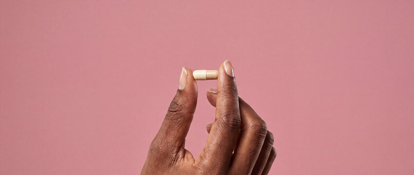 Hand holding a small medical capsule pill in a close-up studio shot against a smooth pink background, ideal for healthcare advertising, pharmaceutical branding, and wellness promotion content