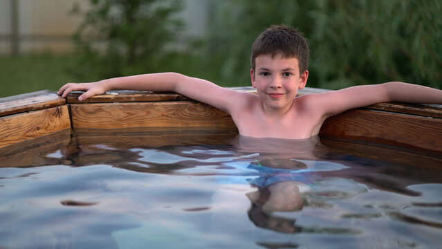 Young boy smiling in outdoor wooden hot tub, arms on rim, relaxing in backyard water at dusk with greenery.