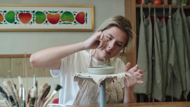Positive girl paints clay plate on special rotating stand. Potters workshop. On background hangs row of aprons - special clothes