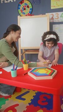 Woman teacher smiling and guiding girl drawing with purple pencil on paper at red activity table in preschool classroom building; nurturing learning.