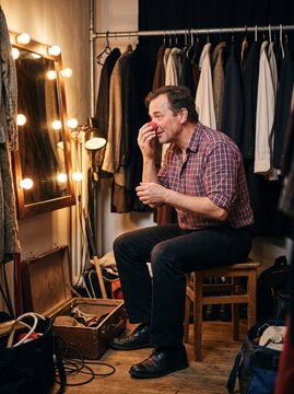 Clown mittleren Alters setzt sich die rote Nase auf und blickt in den Spiegel einer engen Garderobe backstage