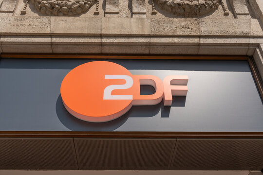 Berlin, Germany July 18, 2026. The orange ZDF television logo is mounted on a dark sign beneath a historic stone facade. Strong sunlight creates bold shadows around the German media emblem.