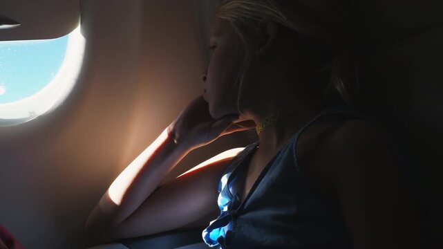 A young girl sits by an airplane window, resting her chin on her hand while looking out at the sky during a flight