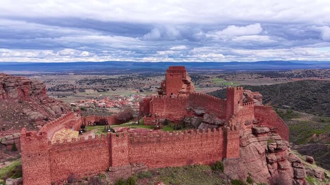 Panoramic view of the medieval Castle of Peracense built on red rock formations in Aragon, Spain, with battlements, tower, catapults and vast landscape under cloudy sky