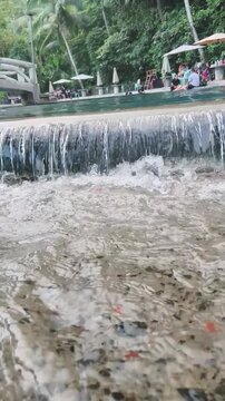 Water cascades over edge into pool with people relaxing nearby under umbrellas