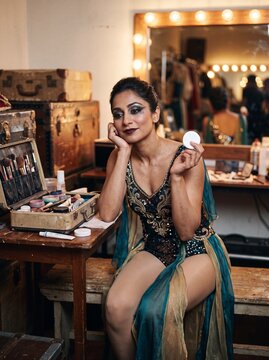 Junge s&uuml;dasiatische Artistin im glitzernden B&uuml;hnenkost&uuml;m sitzt nachdenklich backstage in der Garderobe und h&auml;lt ein Wattepad