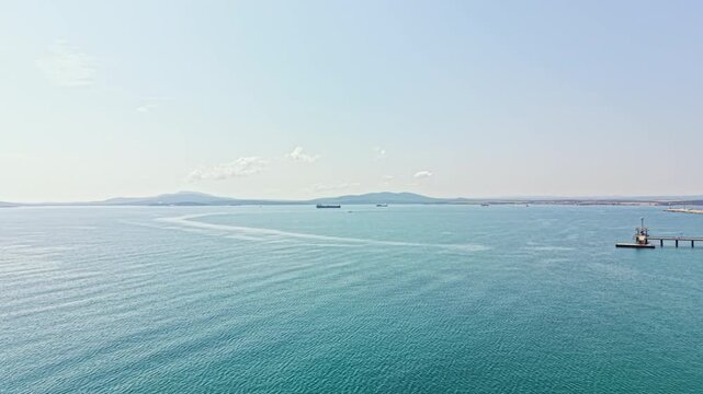 Water stretches across the scene with ships in the distance. A pier stands at the edge where the ocean meets the land. The sky is bright and clear with few clouds.