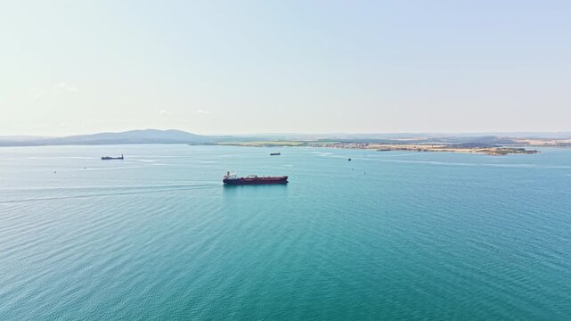 Several cargo ships move slowly through open waters beside a shoreline. The scene shows a bright day with clear visibility and light waves.