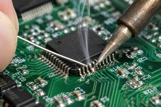 Close-up of a person soldering an integrated circuit on a green PCB with smoke rising from the tip with focus on electronic components.
