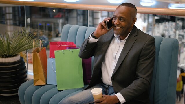 African American mature business man talking phone with coffee cup near shopping bags at mall. Ethnic middle-aged businessman male employer entrepreneur talk mobile call happy communicate hold drink