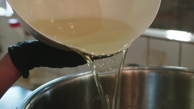 Process of homemade soap. Pouring oil from plastic container into large stainless steel pan. Cooking in the kitchen