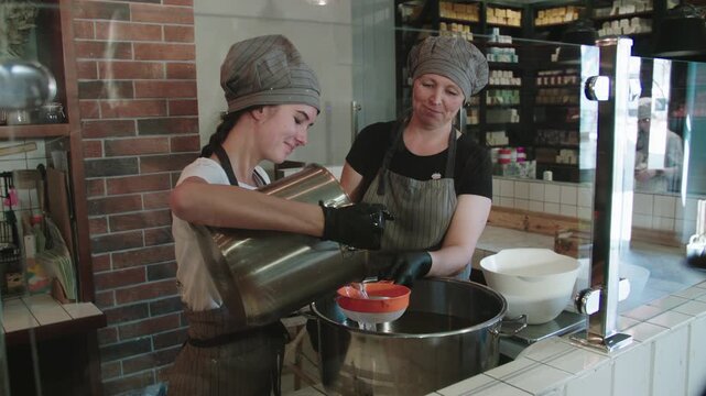 Women in caps, aprons and protective gloves filter alkali solution for pouring it from one container to another through sieve at Craft soap factory.