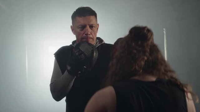 Punching focus mitts held by middle-aged Caucasian male trainer, young Caucasian female boxing. Clip perfect for fitness center marketing and weight loss campaigns