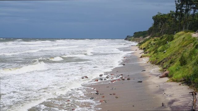 Rough sea waves hitting rocky sandy shore with dense green vegetation under cloudy blue sky