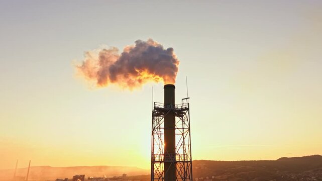 Steam rises from a tall smoke stack in a factory as the sun sets behind the hills. The scene shows industrial activity at dusk, with shadows extending across the landscape.