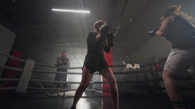 Caucasian young women sparring in boxing ring while male coach watches in background. Footage ideal for fitness brands, female empowerment campaigns, and sports training advertisements