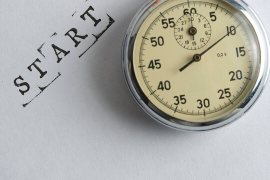 Close up shot an analog stopwatch next to stamped word START on clean white paper surface.