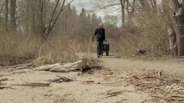 Senior man pulls wheeled utility cart from woodland trail onto sandy beach toward Lake Ontario at Lynde Shores Conservation Area, Whitby, Ontario, Canada.
