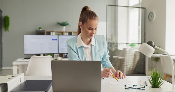 Businesswoman writing documents at laptop in office. She reviews reports, notes findings, and runs analysis at a tidy desk with computer paperwork. Clear business planning and analysis concept