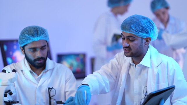Two masked male scientists in caps review a tablet at a bench while colleagues work at a microscope behind under moody blue light in a biotechnology research lab