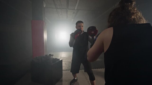 Middle-aged Caucasian male trainer holding focus mitts while young Caucasian female boxer practicing punches in boxing gym. Ideal for fitness center ads and personal training promos