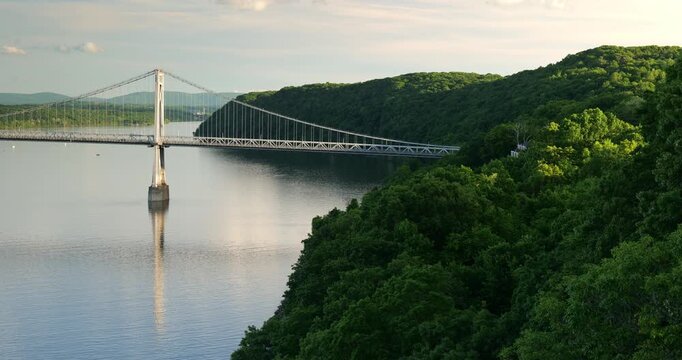 Large suspension bridge spanning across a wide calm Hudson River with forested shores