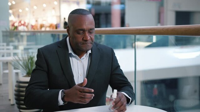 African American mature business man take off medical mask cleaning hands antibacterial spray calling waiter in cafe in shopping mall middle-aged businessman male sanitizing arms coronavirus pandemic