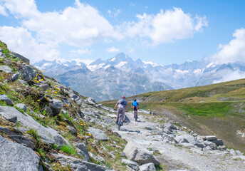 Fototapeta premium Rear view of two unrecognizable mountain bikers cycling along a rugged rocky trail with expansive alpine scenery and distant snow-covered peaks in Switzerland