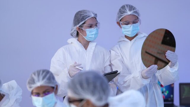 Semiconductor engineers gather around an engineer holding an iridescent silicon wafer with a tablet during a microchip quality inspection in a sterile hitech cleanroom