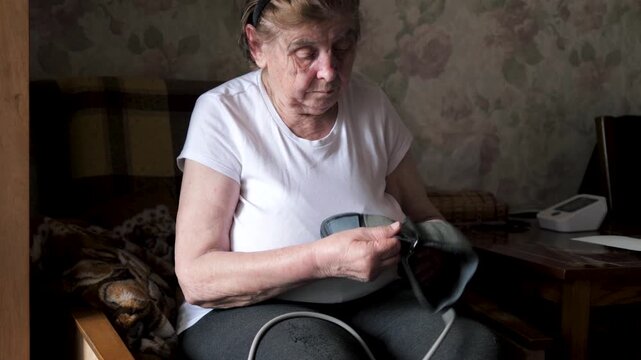 Elderly woman sits at home in armchair and removes cuff of automatic blood pressure monitor from hand and puts it on table. Checking blood pressure and pulse. Attention to health and care of elderly.