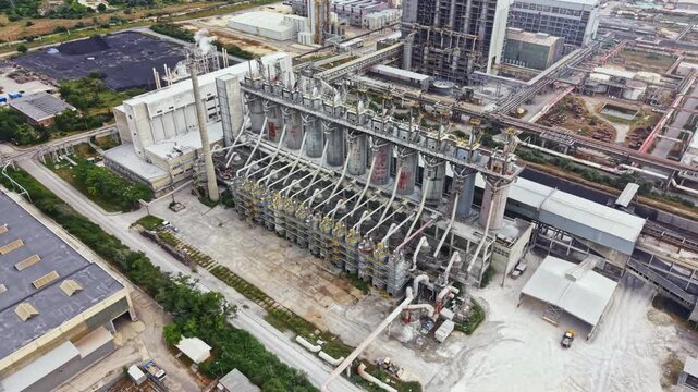 A large industrial site features multiple tall structures with pipes and machinery in operation. The area is busy with activity, showing different sections of the facility.