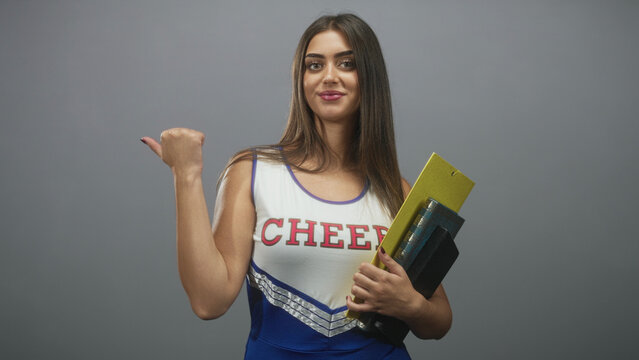 Cheerleader woman holding a yellow folder and book, thumb pointing back gesture while smiling and standing in studio; confidence.