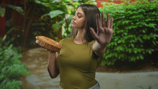 Woman holding cake base with left hand while right hand palms outward stop gesture in forest; defiance.