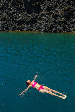 Woman floating in turquoise water near the volcanic cliffs of Santorini, Greece, at the popular hot springs and swimming area visited by catamaran and sailing tours.