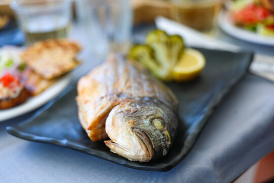 Fresh grilled whole fish served on a black plate with lemon and vegetables at a seaside restaurant in Santorini, showcasing traditional Greek Mediterranean cuisine.