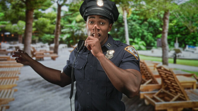 Man in police uniform with visible badge and radio microphone, finger to lips for silence while smiling and pointing arm in street park setting; calm authority.
