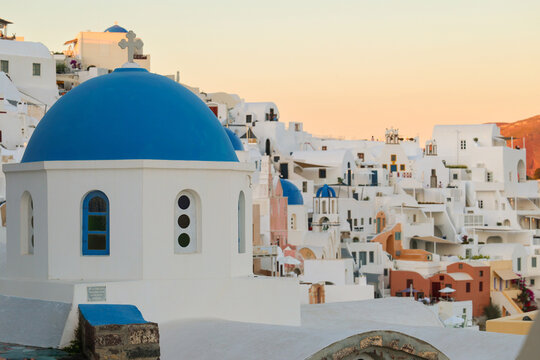 Iconic blue dome church overlooking the whitewashed village of Oia, Santorini, Greece, during golden hour with classic Cycladic architecture and hillside views.