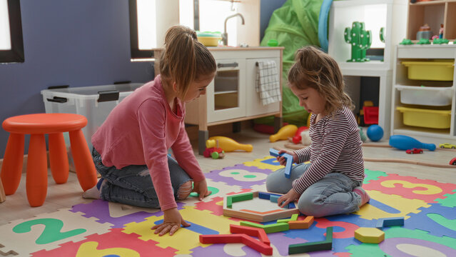 Two little girls kneel on a colorful foam number mat in a building playroom, holding and stacking wooden geometric blocks; playful learning friendship.