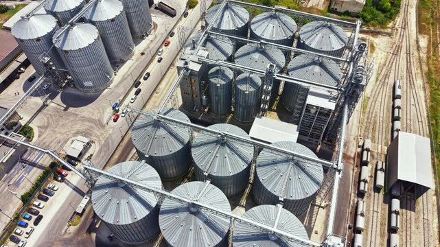 A view shows multiple silos storing grains and a train passing by on a railway nearby. The industrial area has vehicles parked in the lot and tracks in the background.