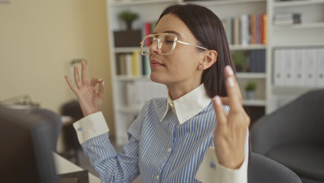Woman at office desk with hands forming ok sign in meditative pose while seated by computer, wearing glasses and striped shirt; mindfulness calm.