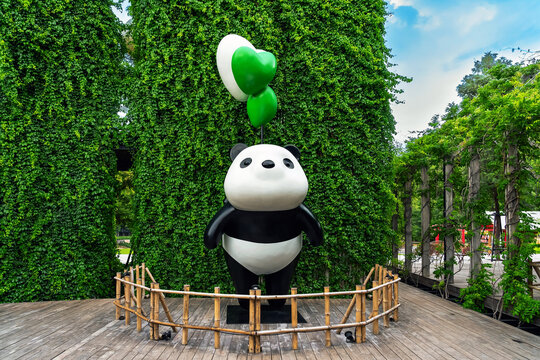 KANCHANABURI, THAILAND - JUNE 12, 2026: A large cute giant panda statue holding green heart balloons on wooden deck with lush green ivy wall background at CHAN nature cafe (local tourist attraction).