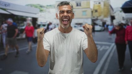 Fototapeta premium Man in white t shirt with fists raised, open mouth cheering and visible forearms on a busy pedestrian street; celebration joy.