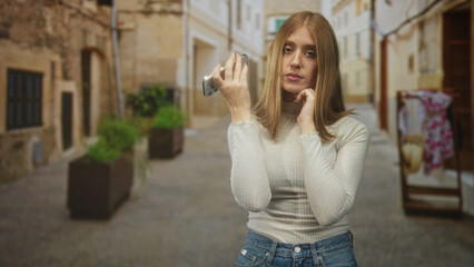 Fototapeta premium Young redhead woman holding smartphone near chin, hand raised, posing on narrow cobblestone street; quiet contemplation.