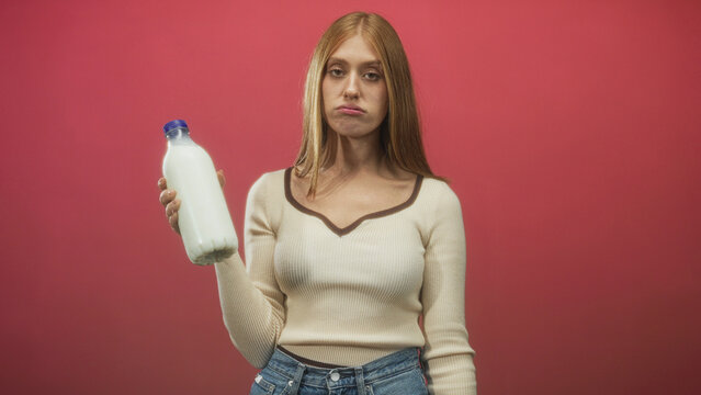 Redhead woman holds a white milk bottle in one hand, puckering lips and rolling eyes while wearing a cream sweater and jeans in a studio red backdrop; skepticism taste testing.
